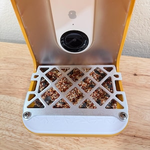 May include: A yellow bird feeder with a built-in camera and a metal feeding tray filled with birdseed. The feeder has a white base and a clear plastic cover. The camera is centered above the tray.