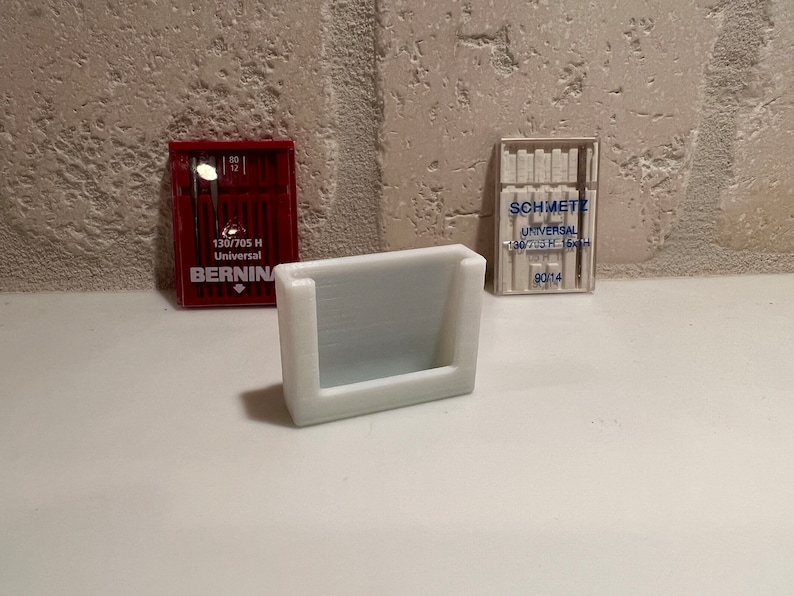 May include: A white plastic needle holder with two packs of sewing machine needles. One pack is red and says "BERNINA 130/705 H Universal". The other pack is clear and says "SCHMETZ UNIVERSAL 130/705 H 13x4".