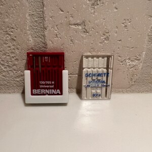 May include: Two packs of sewing machine needles. The pack on the left is red and white and says "130/705 H Universal BERNINA". The pack on the right is clear and white and says "SCHMETZ UNIVERSAL 130/705 H - 15x1H 90/14".