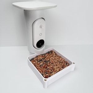 May include: A white bird feeder with a built-in camera. The feeder has a square seed tray filled with birdseed and a camera unit with a lens. The feeder is mounted on a white surface.