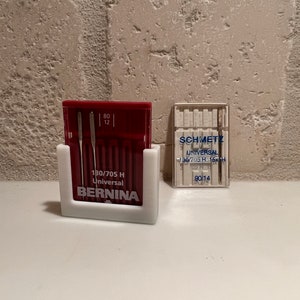 May include: A white plastic sewing needle holder with a red and white box of 12 sewing needles labeled "80/705 H Universal BERNINA". A clear plastic box of sewing needles labeled "SCHMETZ UNIVERSAL 130/705 H-15xH 90/14" is to the right.