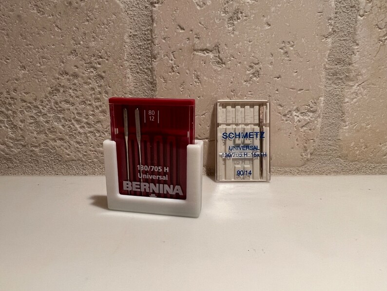 May include: A white plastic sewing needle holder with a red and white box of 12 sewing needles labeled "80/705 H Universal BERNINA". A clear plastic box of sewing needles labeled "SCHMETZ UNIVERSAL 130/705 H-15xH 90/14" is to the right.