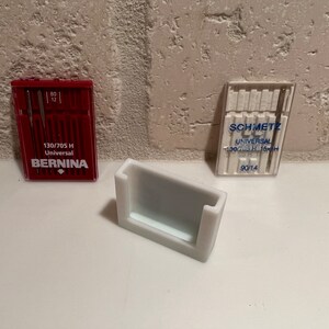 May include: Two packs of sewing machine needles, one red and one white, with the brand names "BERNINA" and "SCHMETZ" printed on them. The packs are sitting on a white surface next to a white plastic needle holder.