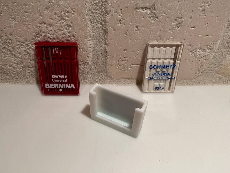 May include: Two packs of sewing machine needles, one red and one white, with the brand names "BERNINA" and "SCHMETZ" printed on them. The packs are sitting on a white surface next to a white plastic needle holder.