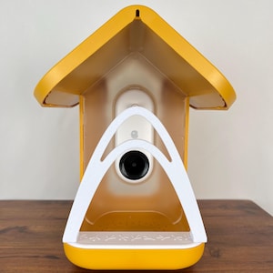 Könnte beinhalten: Ein gelbes Vogelhaus mit einem weißen, bogenförmigen Griff und einem schwarzen Kameraobjektiv. Das Vogelhaus hat ein Dach und eine kleine Sitzstange mit Vogelsilhouetten. Das Vogelhaus ist aus Kunststoff und steht auf einer Holzoberfläche.