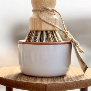 May include: A white ceramic dish with a brown rim holds a wooden handled brush with natural bristles. The brush is tied with twine and the dish is sitting on a wooden table.