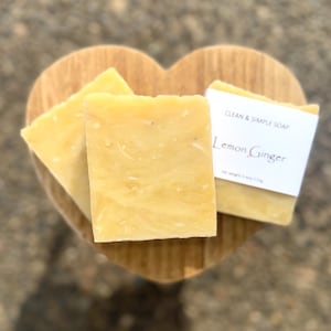 May include: Three bars of yellow soap with a marbled texture, resting on a heart-shaped wooden dish. One bar has a white label that reads "Clean & Simple Soap" and "Lemon Ginger".
