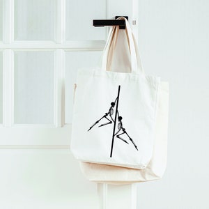 May include: A white canvas tote bag with a black graphic of two skeletons holding onto a pole. The bag is hanging on a white door.