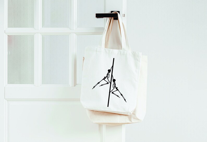 May include: A white canvas tote bag with a black graphic of two skeletons holding onto a pole. The bag is hanging on a white door.