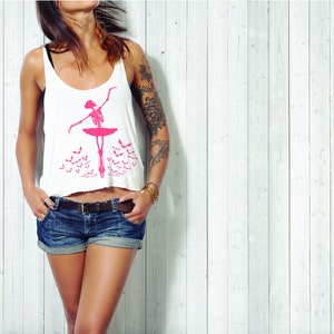 May include: White tank top with a pink skeleton ballerina graphic. The ballerina is posed en pointe on a pink platform, with a pink tutu. Below the ballerina are several pink bat silhouettes. The top is paired with denim shorts.