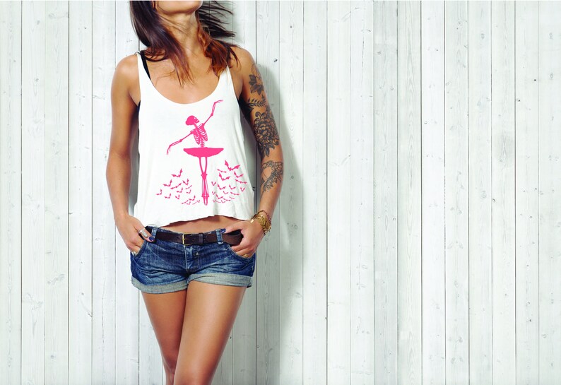May include: White tank top with a pink skeleton ballerina graphic. The ballerina is posed en pointe on a pink platform, with a pink tutu. Below the ballerina are several pink bat silhouettes. The top is paired with denim shorts.