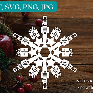 May include: White paper snowflake design featuring a nutcracker and gift boxes. The snowflake is on a brown wooden background. "Nutcracker Snowflake" is written below the snowflake.