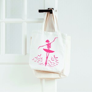 May include: Two off-white canvas tote bags hanging on a black door handle. The front bag features a pink skeleton ballerina design with a tutu and bats. The bags are a light beige color and are hanging on a white door.