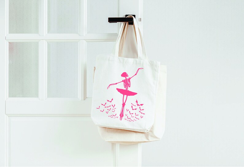 May include: Two off-white canvas tote bags hanging on a black door handle. The front bag features a pink skeleton ballerina design with a tutu and bats. The bags are a light beige color and are hanging on a white door.