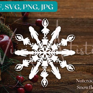 Puede incluir: Silueta blanca de un copo de nieve con un diseño de bailarina. El copo de nieve está formado por seis puntas con una figura de bailarina en cada punta. El copo de nieve está sobre un fondo de madera marrón. El texto "Nutcracker Snowflake" está debajo del copo de nieve.