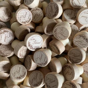 Può includere: Una collezione di tappi per bottiglie in legno. Ogni tappo ha una finitura in legno chiaro e una base in sughero naturale. Le cime sono incise con i nomi "Léa & Thibaud" e la data "15 Juin 2024" circondati da un motivo floreale.