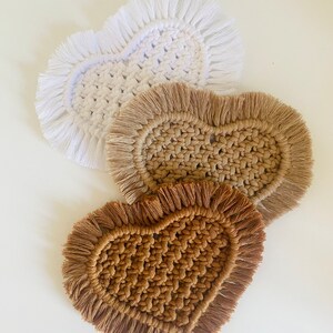 Peut inclure: Trois dessous de verre en macramé en forme de cœur, blancs, beiges et bruns. Chaque dessous de verre présente un centre tissé et un bord à franges. Les dessous de verre sont disposés sur une surface blanche, mettant en valeur leur texture et leur design faits à la main.