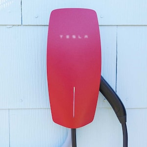 May include: A red Tesla wall connector mounted on a light blue wall. The charger has a sleek, modern design with the Tesla logo visible at the top. A black charging cable is connected to the unit.