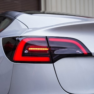 May include: A close-up of the rear taillight of a silver Tesla Model 3. The taillight is a dark smoked lens with a red LED strip.