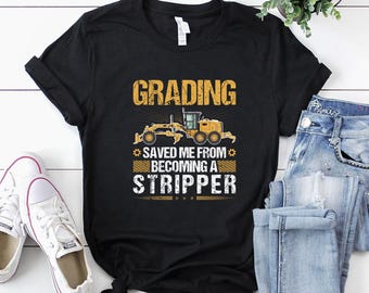 May include: Black t-shirt with the text "GRADING SAVED ME FROM BECOMING A STRIPPER" in yellow and white. The shirt features a yellow construction vehicle graphic. White sneakers and blue jeans are also in the image.