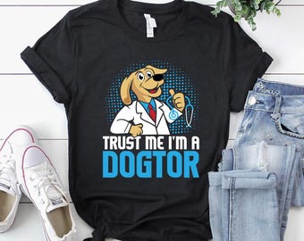 May include: Black t-shirt featuring a cartoon dog dressed as a doctor, with the text "TRUST ME I'M A DOGTOR" in blue and white. The dog is giving a thumbs up and holding a stethoscope. The shirt is paired with white sneakers and blue jeans.