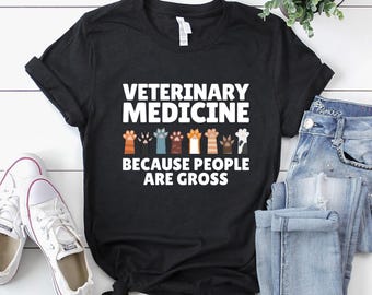 May include: Black t-shirt with the text "VETERINARY MEDICINE BECAUSE PEOPLE ARE GROSS" in white. The design includes colorful paw prints. The shirt is paired with white sneakers and blue jeans.