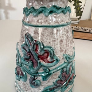 May include: A ceramic vase with a white base and green and red floral designs. The vase has a wide mouth and a narrow base.