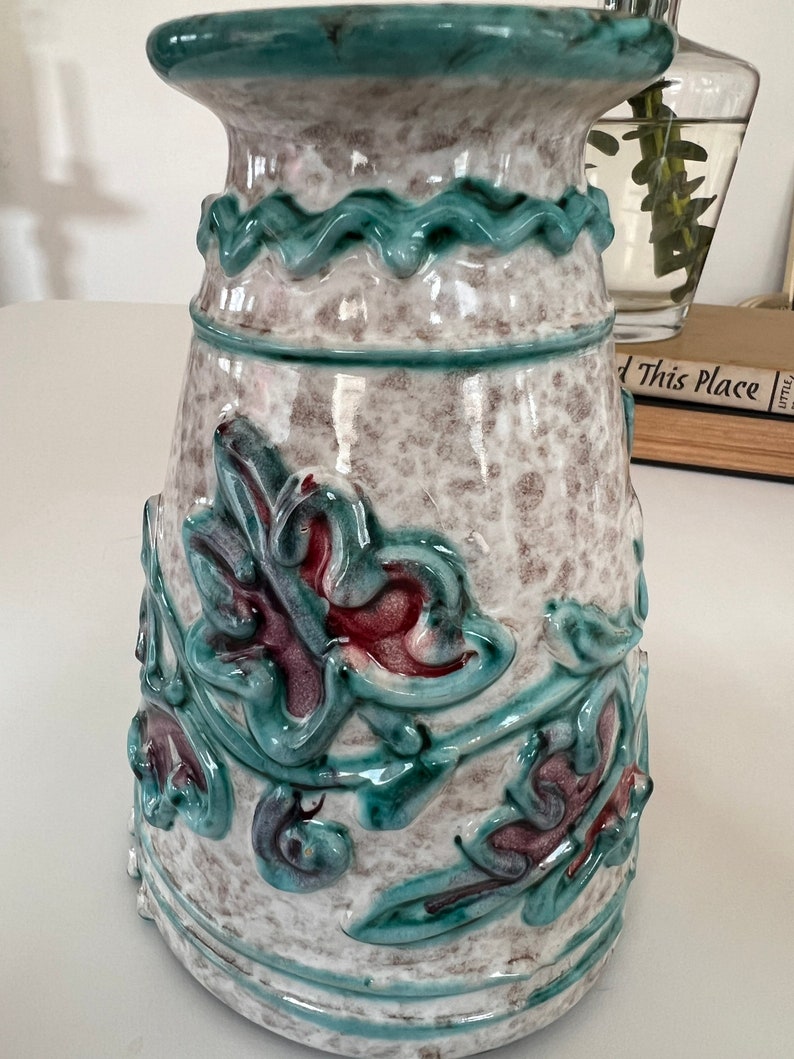 May include: A ceramic vase with a white base and green and red floral designs. The vase has a wide mouth and a narrow base.