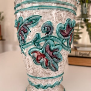 May include: A white ceramic vase with a teal and purple floral design. The vase has a textured surface and a scalloped rim.