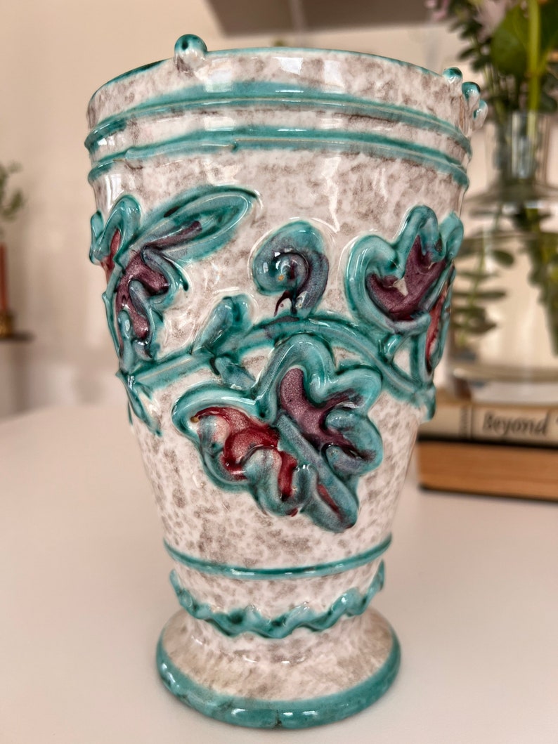 May include: A white ceramic vase with a teal and purple floral design. The vase has a textured surface and a scalloped rim.