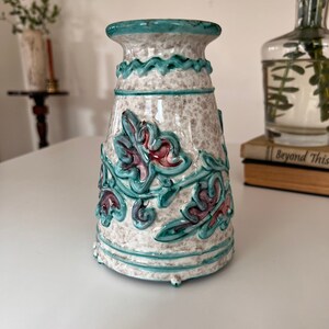 May include: A white ceramic vase with a teal blue rim and floral design. The vase has a textured surface and a raised floral pattern in teal blue, pink, and white.