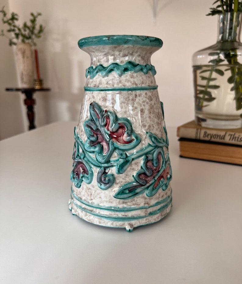 May include: A white ceramic vase with a teal blue rim and floral design. The vase has a textured surface and a raised floral pattern in teal blue, pink, and white.