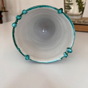 May include: A white ceramic vase with a teal green rim and decorative knobs. The vase is empty and has a smooth, glossy finish.