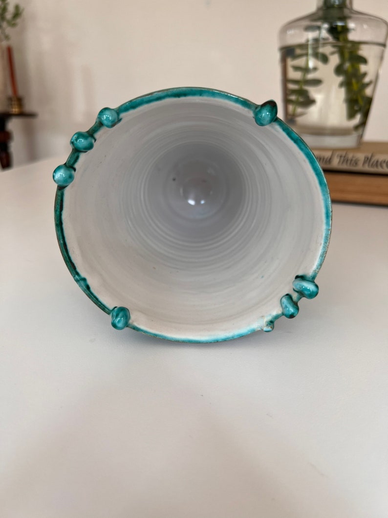 May include: A white ceramic vase with a teal green rim and decorative knobs. The vase is empty and has a smooth, glossy finish.