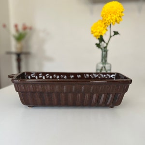May include: A brown ceramic rectangular baking dish with a white glaze and a handle on one side. The dish has a ribbed design and is sitting on a white surface.