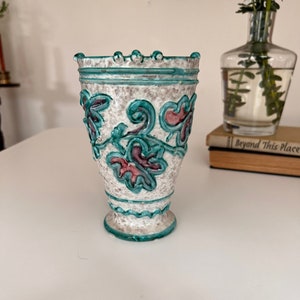 May include: A white ceramic vase with a teal and pink floral design. The vase has a textured surface and a scalloped rim.