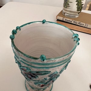 May include: A white ceramic vase with a green and white floral design. The vase has a wide mouth and a round base.