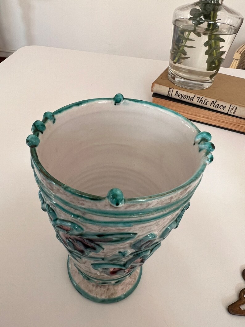 May include: A white ceramic vase with a green and white floral design. The vase has a wide mouth and a round base.