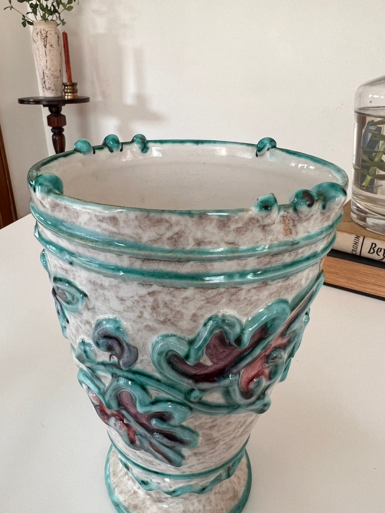 May include: A white ceramic vase with a green and purple floral design. The vase has a wide mouth and a narrow base.