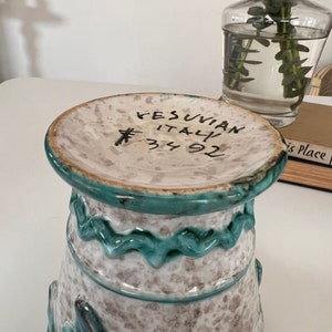 May include: A white ceramic vase with a green glaze and a floral design. The vase has a wide mouth and a narrow base. The vase is marked "VESUVIAN ITALY 34 92".