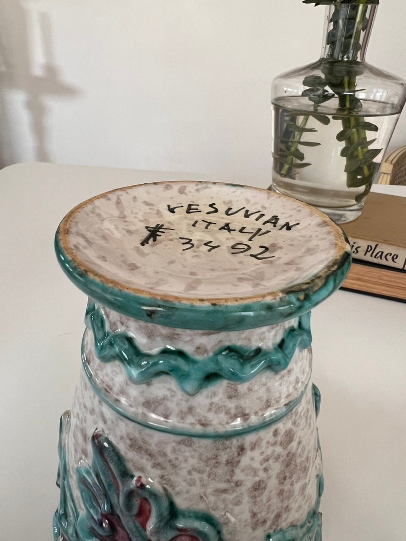 May include: A white ceramic vase with a green glaze and a floral design. The vase has a wide mouth and a narrow base. The vase is marked "VESUVIAN ITALY 34 92".