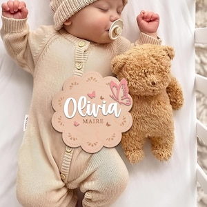 May include: A wooden baby name sign with a pink butterfly and the name "Olivia Maire" in white script. The sign is decorated with a floral pattern and is held by a baby wearing a beige knitted onesie.