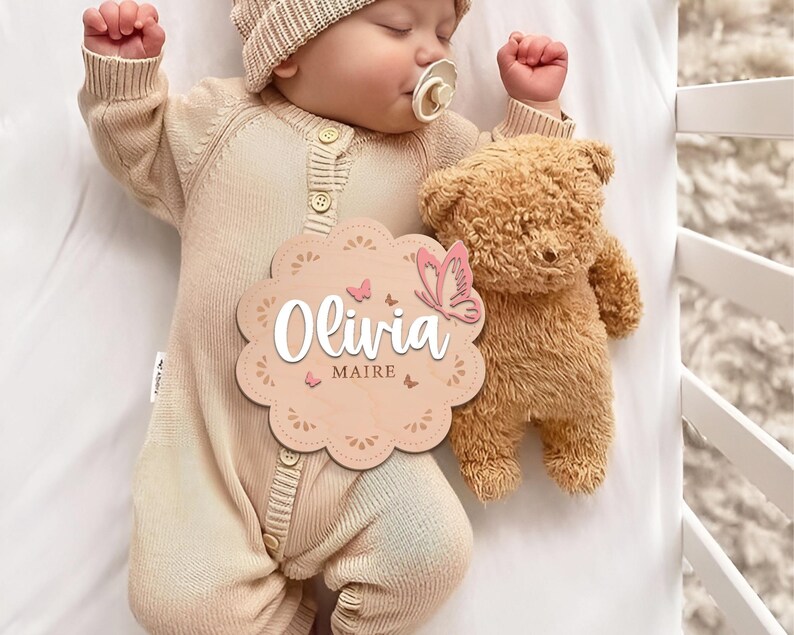 May include: A wooden baby name sign with a pink butterfly and the name "Olivia Maire" in white script. The sign is decorated with a floral pattern and is held by a baby wearing a beige knitted onesie.