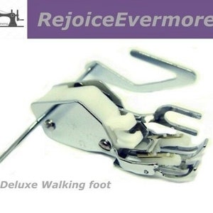 May include: A silver sewing machine walking foot with a white plastic attachment. The foot is designed for use with a sewing machine and is labeled "Deluxe Walking Foot".