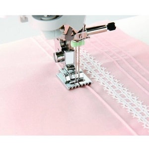 May include: A close-up of a sewing machine foot attached to a sewing machine, sewing a white lace trim to a pink fabric.