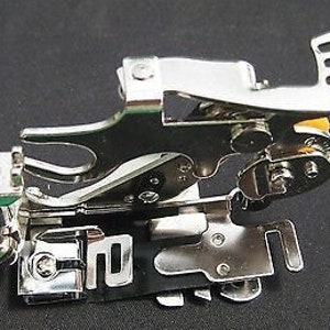 May include: A silver sewing machine foot with a green and silver attachment. The foot has a lever and a series of holes.