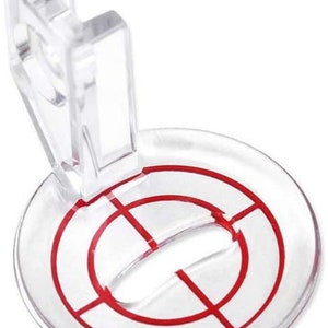 May include: A clear plastic sewing foot with a red target design. The foot has a raised section that can be used to hold fabric in place.
