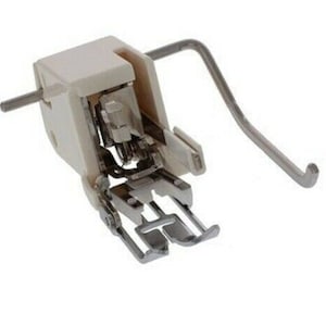 May include: A sewing machine presser foot, featuring a white plastic housing and metallic components. The foot has a rectangular shape with a curved metal arm extending from the side. The design includes a needle opening and a feed dog.