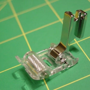 May include: A clear plastic sewing machine foot with a silver metal attachment. The foot has a roller on the bottom.
