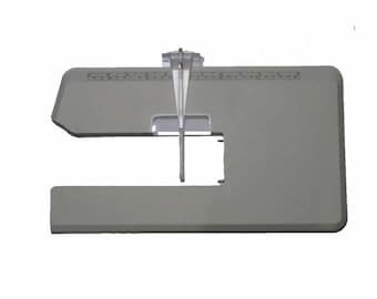 May include: A gray sewing machine extension table with a clear plastic guide and ruler markings. The table has a rectangular shape with a cut-out section and a clear plastic guide that extends downward.